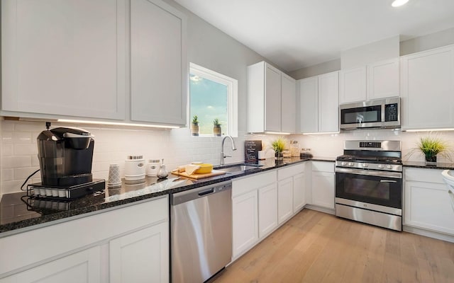 kitchen featuring light wood-type flooring, dark stone countertops, stainless steel appliances, white cabinetry, and a sink