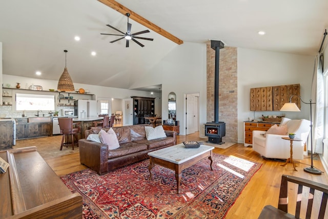 living room with a ceiling fan, a wood stove, light wood-style floors, high vaulted ceiling, and beam ceiling