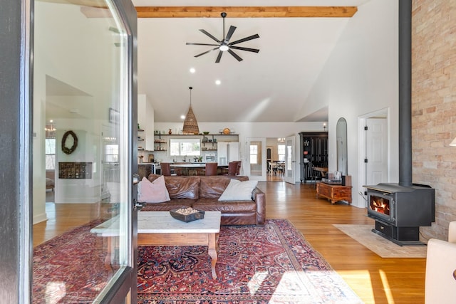 living area with light wood-style flooring, a ceiling fan, a wood stove, high vaulted ceiling, and beamed ceiling