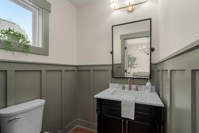 half bath featuring a decorative wall, a wainscoted wall, toilet, a notable chandelier, and vanity