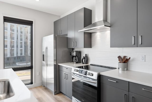 kitchen with decorative backsplash, light wood-style flooring, appliances with stainless steel finishes, gray cabinetry, and wall chimney range hood