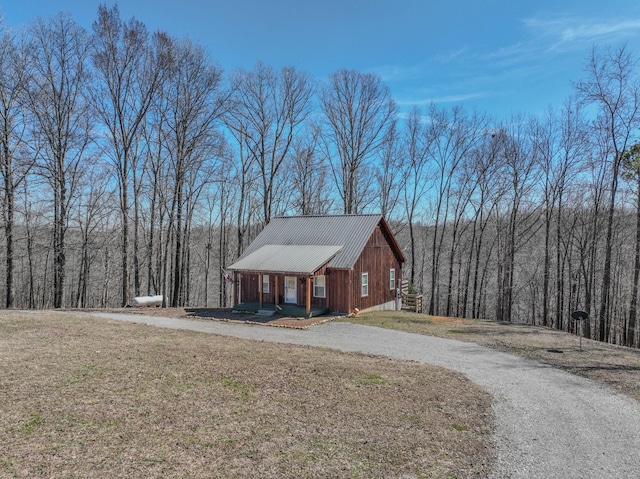 rustic home featuring gravel driveway, a porch, a wooded view, and a front yard