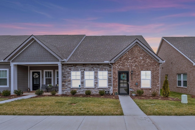 view of front of house featuring stone siding, roof with shingles, a lawn, and brick siding