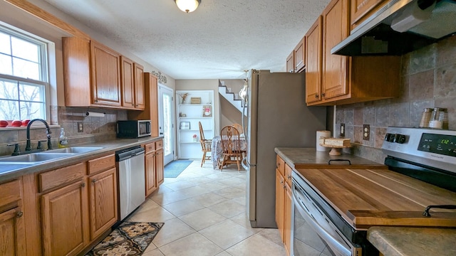 kitchen with brown cabinets, stainless steel appliances, decorative backsplash, a sink, and under cabinet range hood