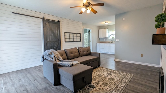 living area featuring a barn door, dark wood-type flooring, ceiling fan, wooden walls, and baseboards