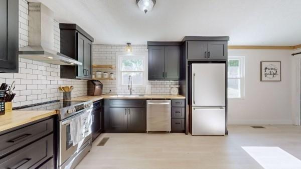 kitchen with stainless steel appliances, a sink, light countertops, wall chimney range hood, and backsplash