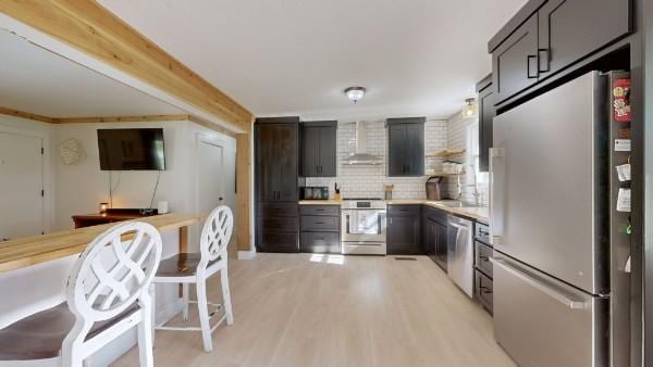 kitchen with light countertops, appliances with stainless steel finishes, light wood-type flooring, decorative backsplash, and wall chimney exhaust hood