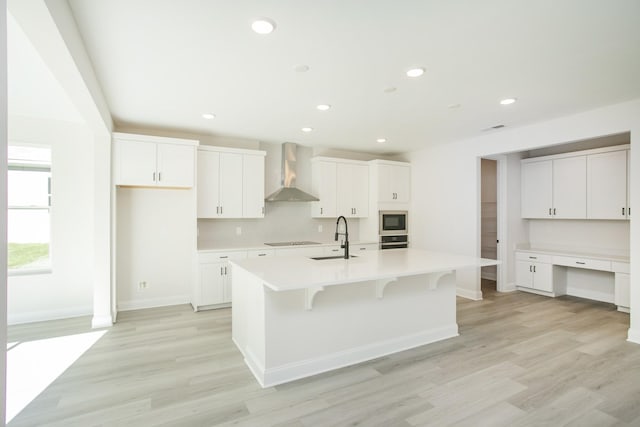 kitchen featuring a center island with sink, appliances with stainless steel finishes, built in study area, a sink, and wall chimney exhaust hood