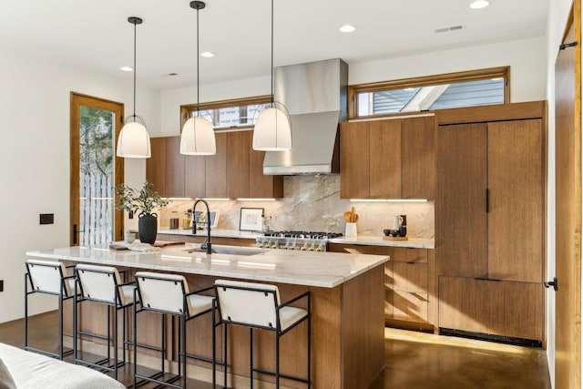 kitchen with wall chimney exhaust hood, brown cabinetry, a sink, and backsplash