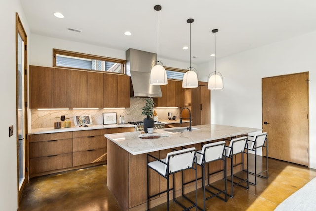 kitchen with tasteful backsplash, wall chimney exhaust hood, brown cabinets, concrete floors, and a sink