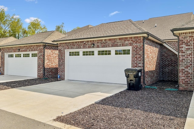 view of side of property with a shingled roof, brick siding, driveway, and a garage