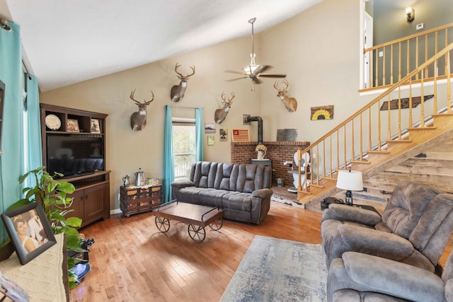 living room with ceiling fan, wood finished floors, a wood stove, stairs, and high vaulted ceiling