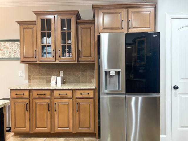 kitchen with light stone countertops, stainless steel refrigerator with ice dispenser, decorative backsplash, brown cabinetry, and glass insert cabinets