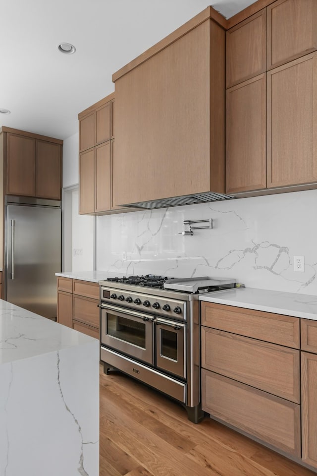 kitchen with high quality appliances, light stone counters, light wood-type flooring, backsplash, and recessed lighting