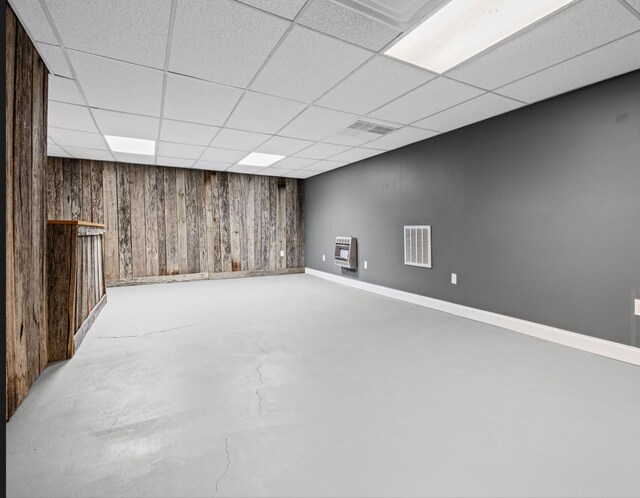 finished below grade area with a paneled ceiling, wooden walls, visible vents, baseboards, and heating unit