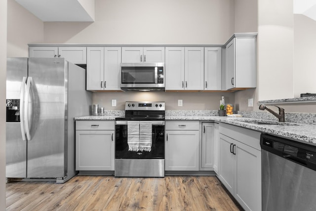 kitchen with stainless steel appliances, gray cabinets, a sink, light stone countertops, and light wood-type flooring