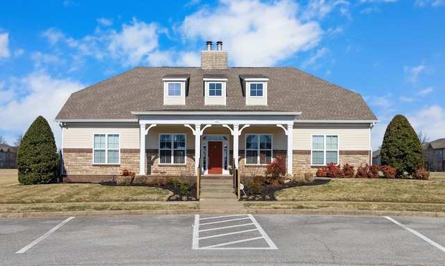 view of front of house with a shingled roof, stone siding, a chimney, covered porch, and a front lawn