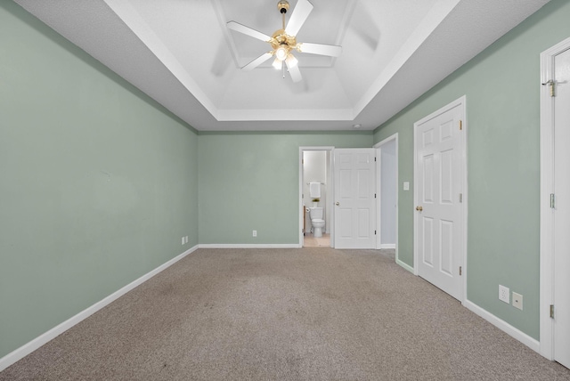 unfurnished bedroom with baseboards, a raised ceiling, ceiling fan, ensuite bathroom, and carpet