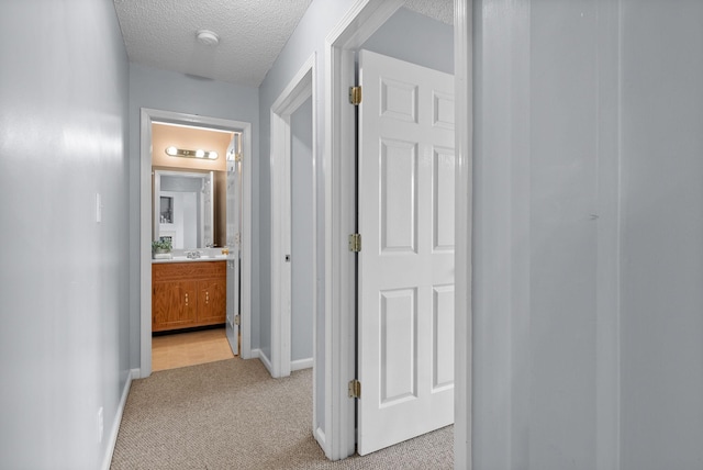 hall with a sink, baseboards, a textured ceiling, and light colored carpet