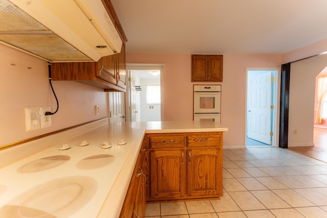 kitchen with brown cabinetry, white double oven, a peninsula, light countertops, and light tile patterned flooring