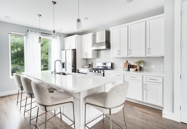 kitchen with an island with sink, dark wood finished floors, stainless steel appliances, wall chimney range hood, and decorative backsplash