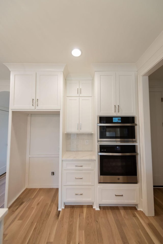 kitchen with tasteful backsplash, white cabinets, light stone counters, light wood-style floors, and double oven