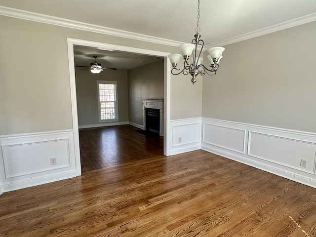 unfurnished dining area featuring ceiling fan with notable chandelier, dark wood-style flooring, a fireplace, ornamental molding, and wainscoting