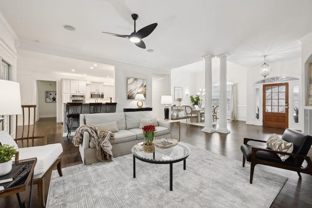 living area with decorative columns, ornamental molding, dark wood-type flooring, and ceiling fan with notable chandelier