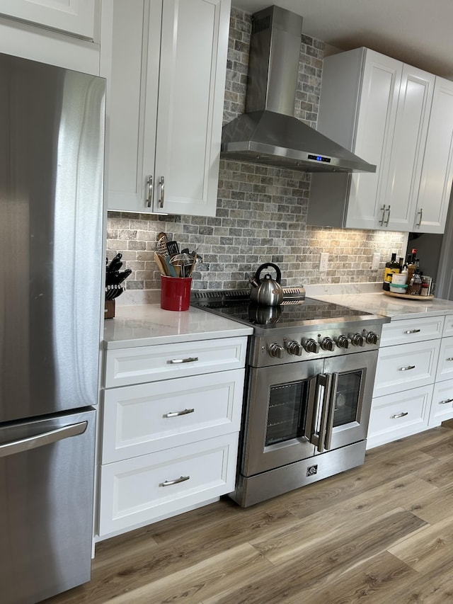 kitchen with wood finished floors, white cabinets, wall chimney range hood, appliances with stainless steel finishes, and backsplash