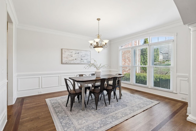 dining space with dark wood-style floors, a wainscoted wall, an inviting chandelier, crown molding, and ornate columns