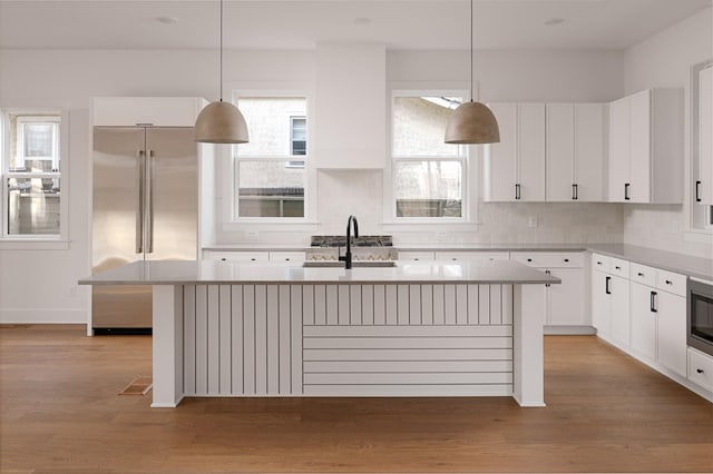 kitchen with light countertops, light wood-style flooring, stainless steel built in fridge, a sink, and a kitchen island with sink