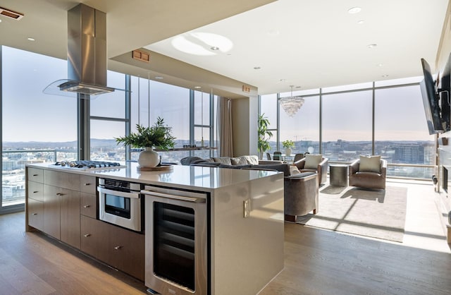 kitchen with beverage cooler, island range hood, appliances with stainless steel finishes, wood finished floors, and a wall of windows