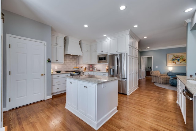 kitchen featuring light wood-style flooring, stainless steel appliances, premium range hood, white cabinetry, and backsplash