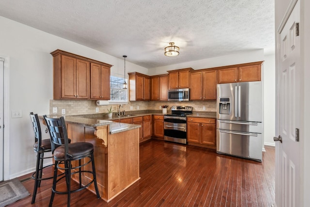 kitchen with dark wood-style floors, a peninsula, brown cabinetry, stainless steel appliances, and a sink