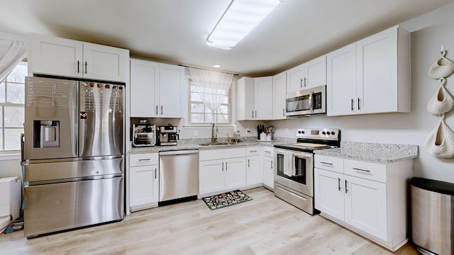 kitchen with white cabinets, appliances with stainless steel finishes, light stone countertops, light wood-style floors, and a sink