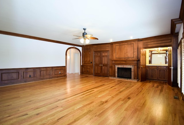 unfurnished living room featuring ceiling fan with notable chandelier, arched walkways, light wood-style floors, crown molding, and a brick fireplace