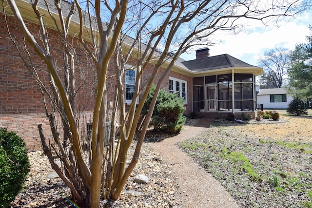 back of house featuring brick siding, a shingled roof, a chimney, and a sunroom