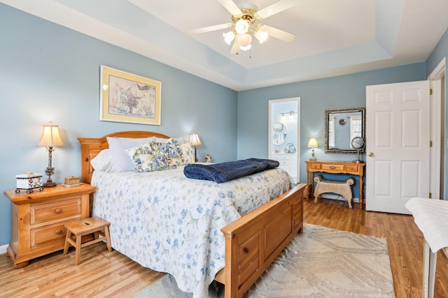 bedroom featuring ceiling fan, a raised ceiling, ensuite bath, and light wood-style flooring