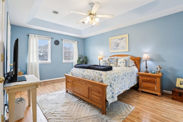 bedroom featuring baseboards, visible vents, light wood finished floors, a tray ceiling, and ceiling fan