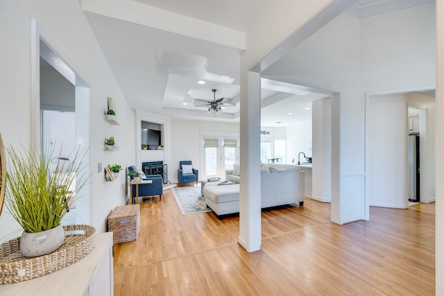 living area featuring light wood-style flooring, a tray ceiling, a glass covered fireplace, recessed lighting, and ceiling fan