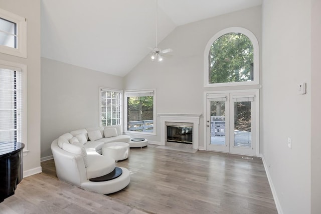 living room featuring wood finished floors, baseboards, high vaulted ceiling, a fireplace with flush hearth, and ceiling fan