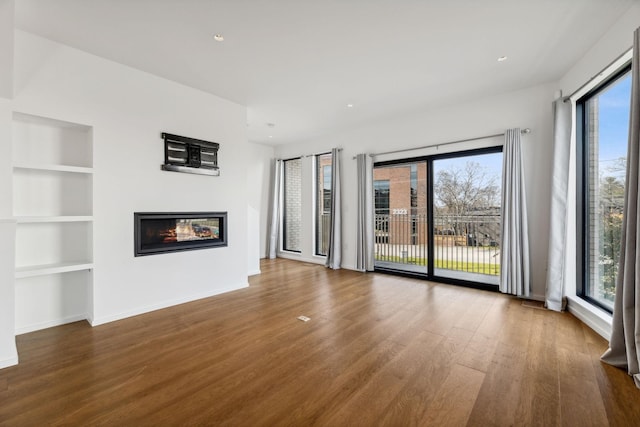 unfurnished living room featuring built in features, wood finished floors, baseboards, recessed lighting, and a glass covered fireplace