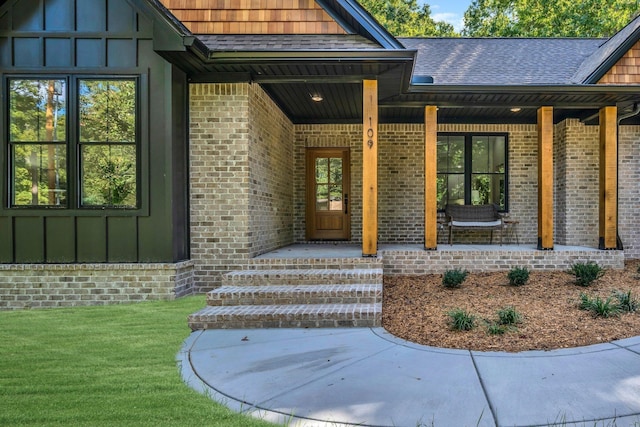 property entrance featuring roof with shingles, a yard, covered porch, board and batten siding, and brick siding