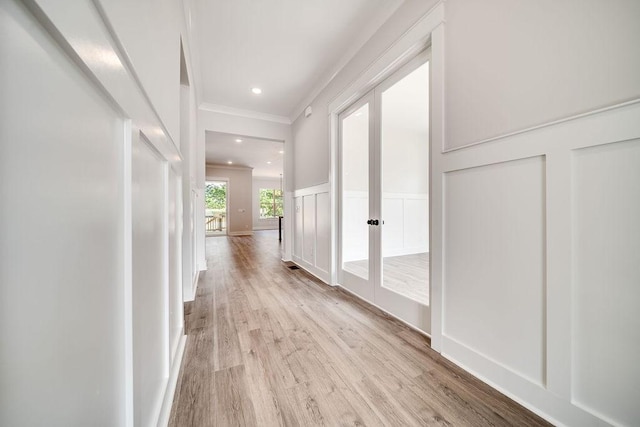 hallway with french doors, a decorative wall, light wood-style floors, wainscoting, and crown molding