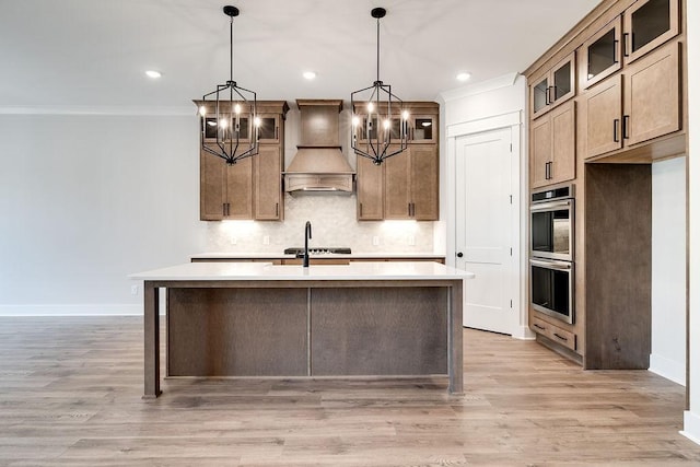 kitchen with light wood-type flooring, a center island with sink, backsplash, light countertops, and custom exhaust hood