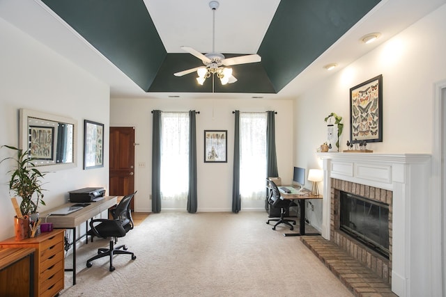 home office with a brick fireplace, baseboards, ceiling fan, light colored carpet, and a tray ceiling