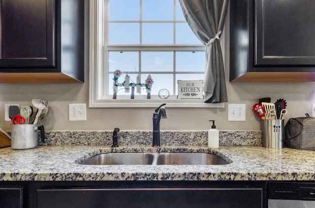 kitchen featuring dark cabinets, light stone countertops, stainless steel dishwasher, and a sink