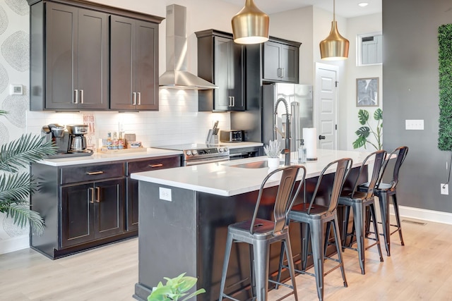 kitchen with wall chimney range hood, decorative backsplash, appliances with stainless steel finishes, a kitchen breakfast bar, and a kitchen island with sink