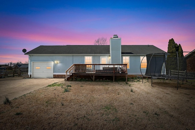 back of property with a wooden deck, an attached garage, a chimney, concrete driveway, and a trampoline