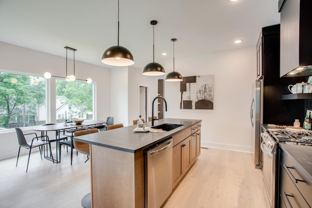 kitchen with light wood finished floors, a kitchen island with sink, a sink, appliances with stainless steel finishes, and dark countertops